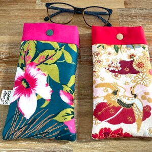 Puede incluir: Dos estuches para gafas de tela con ribetes rosa y rojo. El estuche de la izquierda tiene un patrón floral en tonos verdes, blancos y rosas. El estuche de la derecha tiene un fondo blanco con un patrón floral rojo y dorado y un diseño de grúa. Ambos estuches tienen un cierre de botón.