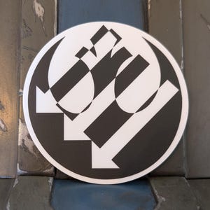 May include: A circular sticker with a black and white graphic design. The design includes diagonal black and white stripes and arrows within a circular border. The sticker is set against a backdrop of weathered wood.