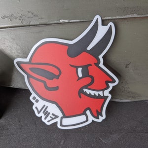 May include: Red and black devil head sticker with a white outline and the word "Jimi" written in black below the head.