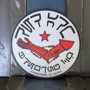Puede incluir: Una pegatina redonda blanca con un diseño de brazo robótico rojo, una estrella roja y texto estilizado. La pegatina tiene un borde negro y un fondo neutro.