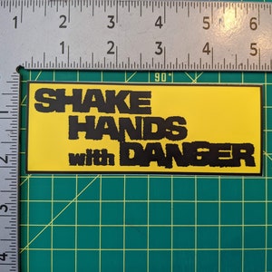 May include: A rectangular sticker with the words "SHAKE HANDS with DANGER" in black, bold, block letters. The background is yellow, and the sticker is outlined in black. The sticker is on a green surface with a ruler.