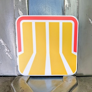 May include: A rectangular sticker with a yellow and white design, outlined in red. The design features three vertical yellow stripes with white borders, creating a stylized architectural or geometric pattern. The sticker is placed on a textured surface.