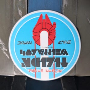 May include: A circular sign featuring a bright blue background with a central red graphic of a spaceship. The design includes white vertical lines and black text in a stylized font, with additional red and black text elements positioned throughout.