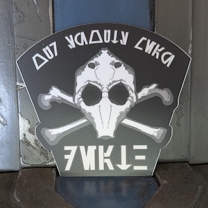 Op de afbeelding: Zwart-witte sticker met een schedel en gekruiste beenderen ontwerp. De schedel draagt ​​een masker en de tekst "DU7 YADUUY CWKA" staat boven de schedel en de tekst "7WKLΞ" staat onder de schedel.