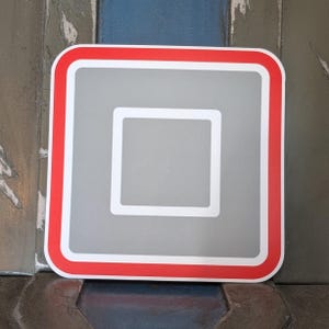 May include: A square sticker with rounded corners. The sticker has a gray center with a white square in the middle. A white border surrounds the gray center, and a red border frames the sticker.