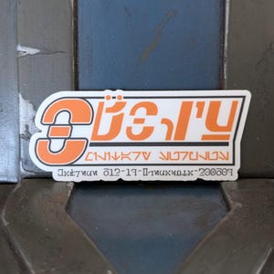 May include: Orange and white sticker with the text "The Mandalorian" in Aurebesh script. The sticker also includes the text "Star Wars" and "Disney+."