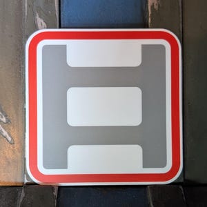 May include: A square sticker with a red border and a gray interior. The sticker features a white, stylized letter "H" shape. The background is a mix of dark gray, blue, and black.