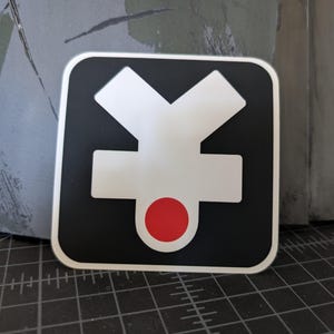 Könnte beinhalten: Ein schwarzer quadratischer Aufkleber mit einem weißen Y-förmigen Symbol und einem roten Kreis in der Mitte.