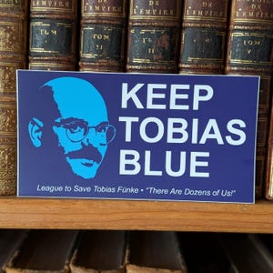 Puede incluir: Una pegatina azul con el texto "KEEP TOBIAS BLUE" y una silueta azul de un hombre con gafas. La pegatina también incluye el texto "League to Save Tobias Fünke. There Are Dozens of Us!"