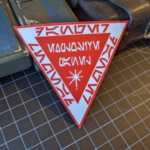 Könnte beinhalten: Ein roter und weißer dreieckiger Aufkleber mit stilisiertem Text und einem Sternmuster. Der Aufkleber hat weißen Text auf rotem Hintergrund, wobei der Text an den Rändern und in der Mitte des Dreiecks angeordnet ist.