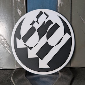May include: A circular emblem featuring a high-contrast black and white graphic design. The pattern consists of bold, intersecting geometric shapes, including sharp diagonal lines and arrow-like forms that create a stylized, abstract composition.