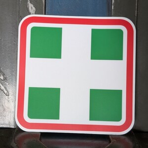 May include: Square sticker with a white background, a red border, and a white cross with four green squares. The sticker has rounded corners and is likely a first aid or medical symbol.