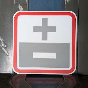 May include: A square sticker with a white background, a red border, and a gray plus sign over a gray minus sign. The sticker is likely used to indicate the positive and negative terminals of a battery or electrical component.