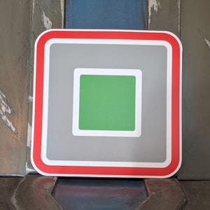 May include: A square sticker with a red border, a white inner border, a gray background, and a green square in the center. The sticker is square with rounded corners.