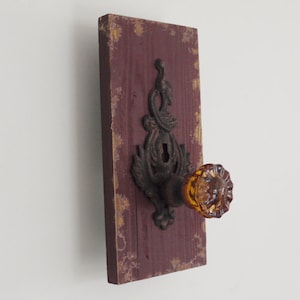 May include: A rustic wooden wall hook with a distressed burgundy finish and gold accents. The hook features a cast iron design with a decorative knob and a glass door knob.