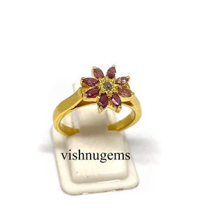 May include: A gold-colored ring featuring a flower design with maroon-colored petals and a small, light-colored center stone. The ring is displayed on a white stand against a white background. The text "vishnugems" is visible.
