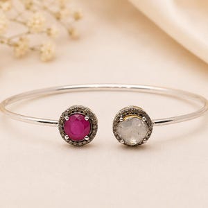 Puede incluir: Una pulsera abierta de plata con dos engastes circulares en los extremos. Un engaste contiene una piedra preciosa redonda de color rosa vibrante, mientras que el otro tiene una piedra blanca translúcida redonda. Ambas rodeadas de pequeños pavés oscuros.
