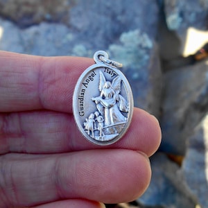 Może przedstawiać: Srebrny owalny wisiorek z motywem anioła stróża. Anioł jest przedstawiony z skrzydłami i trzyma małe dziecko. Wisiorek ma wygrawerowane słowa "Guardian Angel".