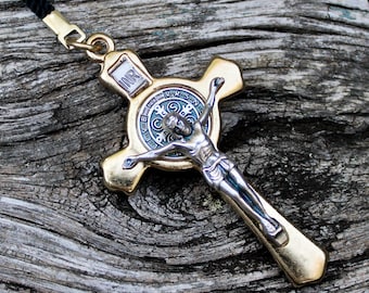 May include: A gold-toned crucifix pendant with a silver Jesus figure and a blue and silver medallion. The cross is suspended from a black cord. The pendant rests on a weathered wooden surface.