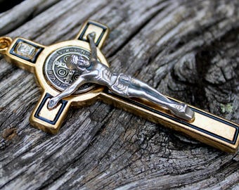 May include: A gold-coloured cross pendant featuring a silver-coloured depiction of Jesus Christ. The cross has black accents and a circular emblem at the centre. The pendant is displayed on a weathered wooden surface.