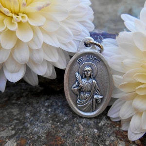 May include: Silver oval pendant with a depiction of Saint Lucy, holding a palm branch and a chalice. The pendant is surrounded by white flowers.