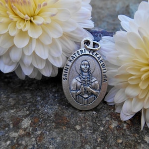 Puede incluir: Pendiente ovalado de plata con una representación de Santa Kateri Tekakwitha, una santa nativa americana, sosteniendo una cruz. El pendiente está rodeado de flores blancas.