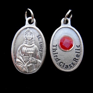 May include: Silver oval pendant with a depiction of Saint Lucy holding a chalice. The back of the pendant has a red relic with the text "Third Class Relic".