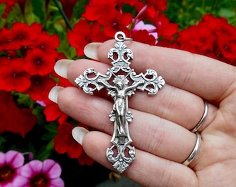 May include: A silver filigree crucifix pendant with a detailed depiction of Jesus Christ on the cross. The pendant is held in a hand against a background of red flowers.