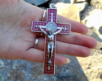 May include: A silver and red crucifix with a depiction of Jesus Christ on the cross. The crucifix has a red enamel background with a white floral pattern and a silver border. The crucifix also features a silver medal with a depiction of Saint Francis of Assisi.