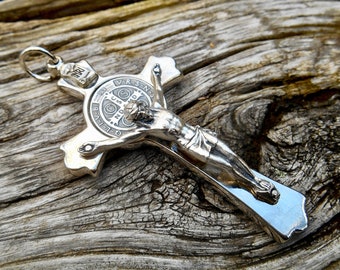 May include: A silver-toned metal crucifix with a figure of Jesus Christ on the cross. The cross features a circular medallion with the inscription "V.R.S.N. - O.L.V.B. - M.D. - J.C. - S.P.D.N. - C.S.S.M. - I.H.S. - I.N.R.I."
