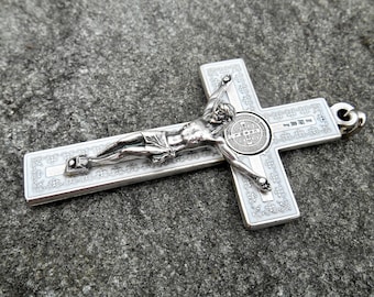 May include: A silver-toned metal crucifix pendant featuring a detailed figure of Jesus attached to the center. The cross has white enamel inlay with intricate decorative patterns and a circular Saint Benedict medal at the intersection point.