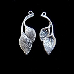 May include: Two silver-colored leaf charms with a textured finish, each featuring three leaves. The charms have a loop at the top for attaching to jewelry. The background is black.