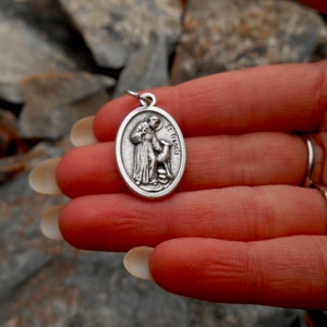 May include: Silver oval pendant with a depiction of Saint Francis of Assisi holding a bird. The pendant has the words "St. Francis" inscribed on it.