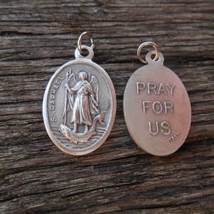 Może przedstawiać: Srebrny, owalny wisiorek z wizerunkiem św. Rafała z jednej strony i napisem "Pray For Us" z drugiej strony. Wisiorek został wykonany we Włoszech.