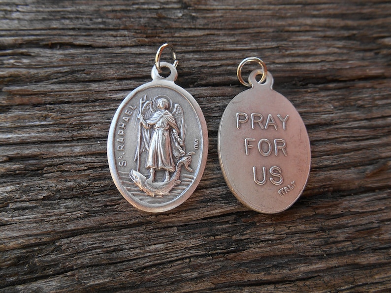 Może przedstawiać: Srebrny, owalny wisiorek z wizerunkiem św. Rafała z jednej strony i napisem "Pray For Us" z drugiej strony. Wisiorek został wykonany we Włoszech.