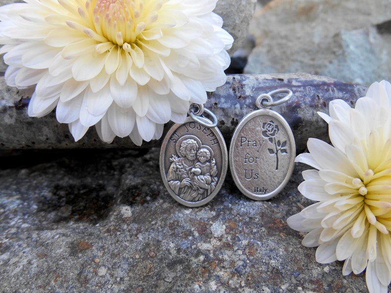 May include: Two silver oval-shaped religious medallions. The first medallion features a depiction of Saint Joseph holding the baby Jesus. The second medallion has the text "Pray for Us" and a rose. Both medallions are on a background of white flowers and gray rocks.