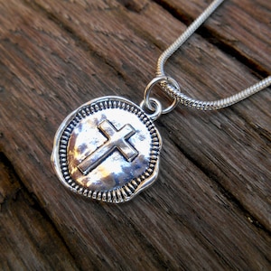 Op de afbeelding: Een zilveren ketting met een ronde hanger met een reliëf van een kruis. De hanger heeft een getextureerde, gehammerde afwerking en een parelrand.