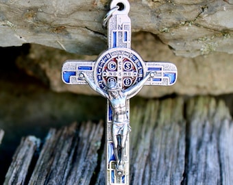 May include: A silver-colored religious cross pendant with a blue and red enamel design. The cross features a detailed depiction of Jesus Christ and is suspended from a small loop. The background is a weathered wooden surface.