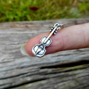 Puede incluir: Un charm de plata con forma de violín. El charm es pequeño y detallado, con un bucle de plata en la parte superior para sujetarlo a una pulsera o collar.
