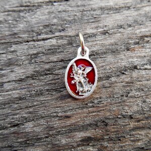 May include: A silver charm with a red enamel background depicting an angel with wings outstretched. The charm is oval shaped and has a loop at the top for attaching to a chain or bracelet.