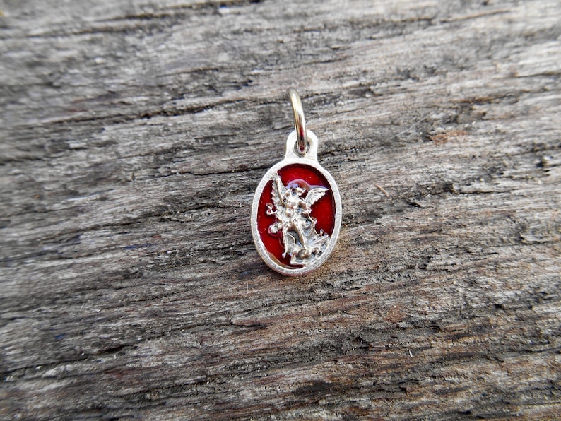 May include: A silver charm with a red enamel background depicting an angel with wings outstretched. The charm is oval shaped and has a loop at the top for attaching to a chain or bracelet.