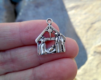 May include: A small, silver-colored charm depicting a nativity scene. The charm features a star, a stable, and figures representing Mary, Joseph, and baby Jesus. The charm has a loop at the top for attaching to a necklace or bracelet.
