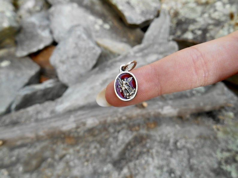 May include: A silver charm with a red enamel background featuring a gold-colored angel with wings. The charm is oval-shaped and has a loop at the top for attaching to a chain.