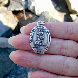 May include: An oval silver-toned metal pendant featuring a raised relief of Saint Joseph holding the Child Jesus. The border is inscribed with the text St. Joseph, Terror of Demons, Pray For Us. A small metal jump ring is attached to the top.