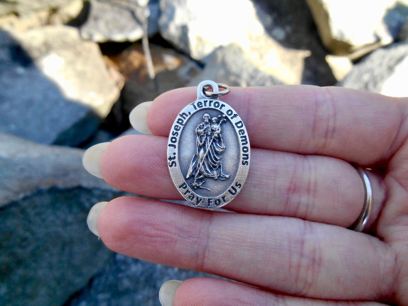 May include: An oval silver-toned metal pendant featuring a raised relief of Saint Joseph holding the Child Jesus. The border is inscribed with the text St. Joseph, Terror of Demons, Pray For Us. A small metal jump ring is attached to the top.