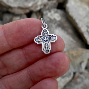 Puede incluir: Pendiente de plata en forma de cruz con cinco peque&ntilde;as figuras religiosas a cada lado. La cruz est&aacute; detallada con un acabado texturizado.