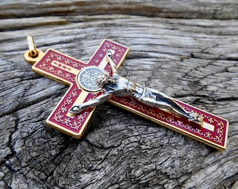 May include: A gold crucifix pendant with a red enamel cross and a silver figure of Jesus. The cross has gold detailing and a circular emblem. The pendant is displayed on a weathered wooden surface.