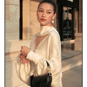 May include: A woman wearing a cream-colored blouse with a high neckline and long sleeves, paired with a black skirt. She carries a black saddlebag with a gold-tone 'D' charm. The background features a building facade.