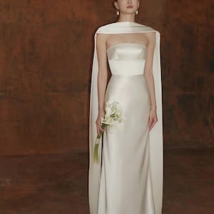 May include: A white strapless wedding gown with a flowing cape. The dress features a fitted bodice and a long, elegant skirt. The bride is holding a bouquet of white flowers. The background is a warm, brown tone.
