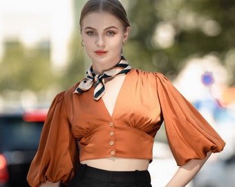 May include: A woman wearing a rust-colored blouse with puffed sleeves and a black and white patterned scarf. She is also wearing black pants and a black clutch.